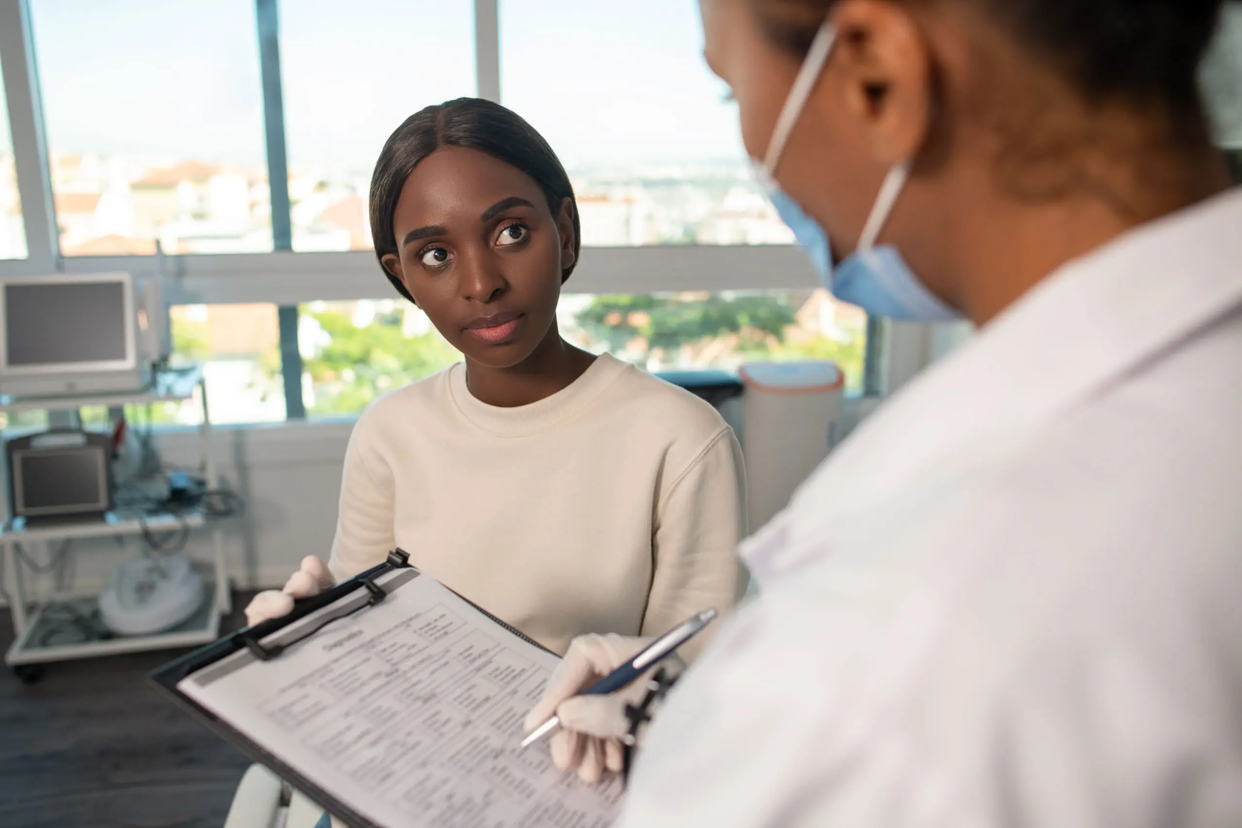 Serious African American woman answering questions of doctor Serious African American woman answering questions of doctor in clinic. General practitioner filling in medical survey of female patient. Medical exam concept