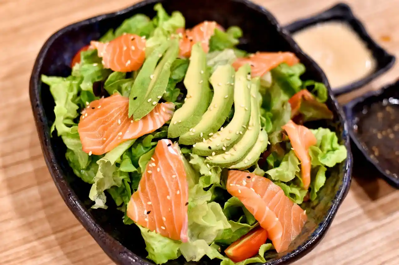 qt=q_30 (4) A salad bowl featuring lettuce, sliced avocado, and raw salmon, garnished with sesame seeds. Two small dishes on the side contain soy sauce and dressing.