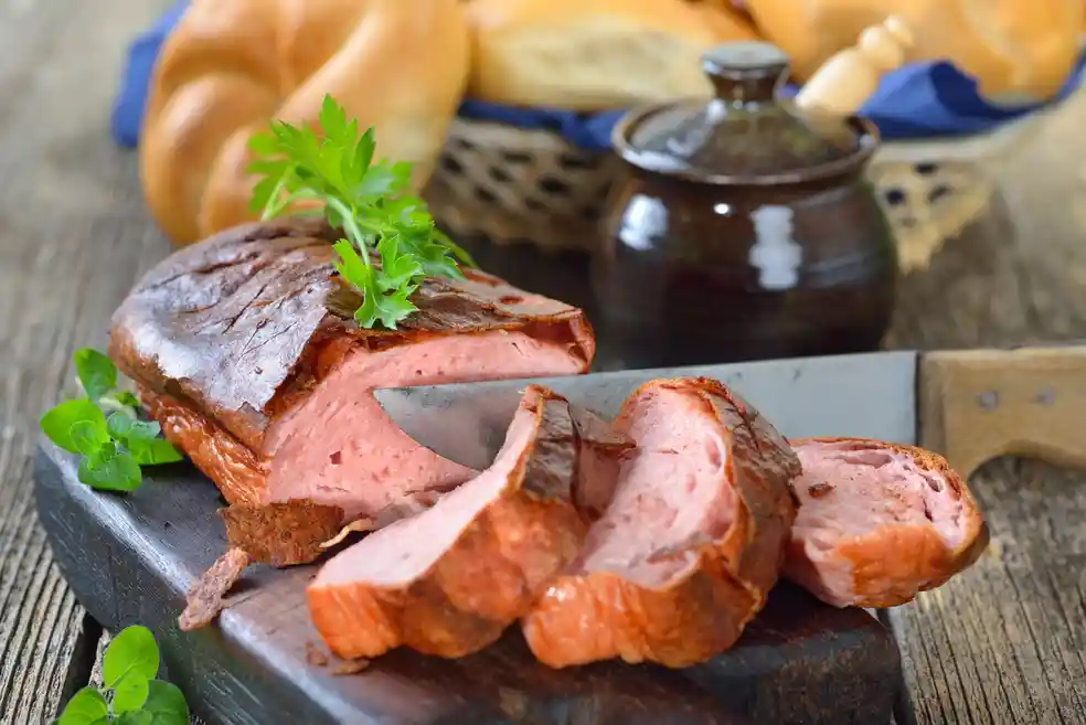 qt=q_30 (5) Sliced pink sausage loaf on a wooden board with parsley garnish. Knife next to slices. Bread basket and sauce pot in the background.