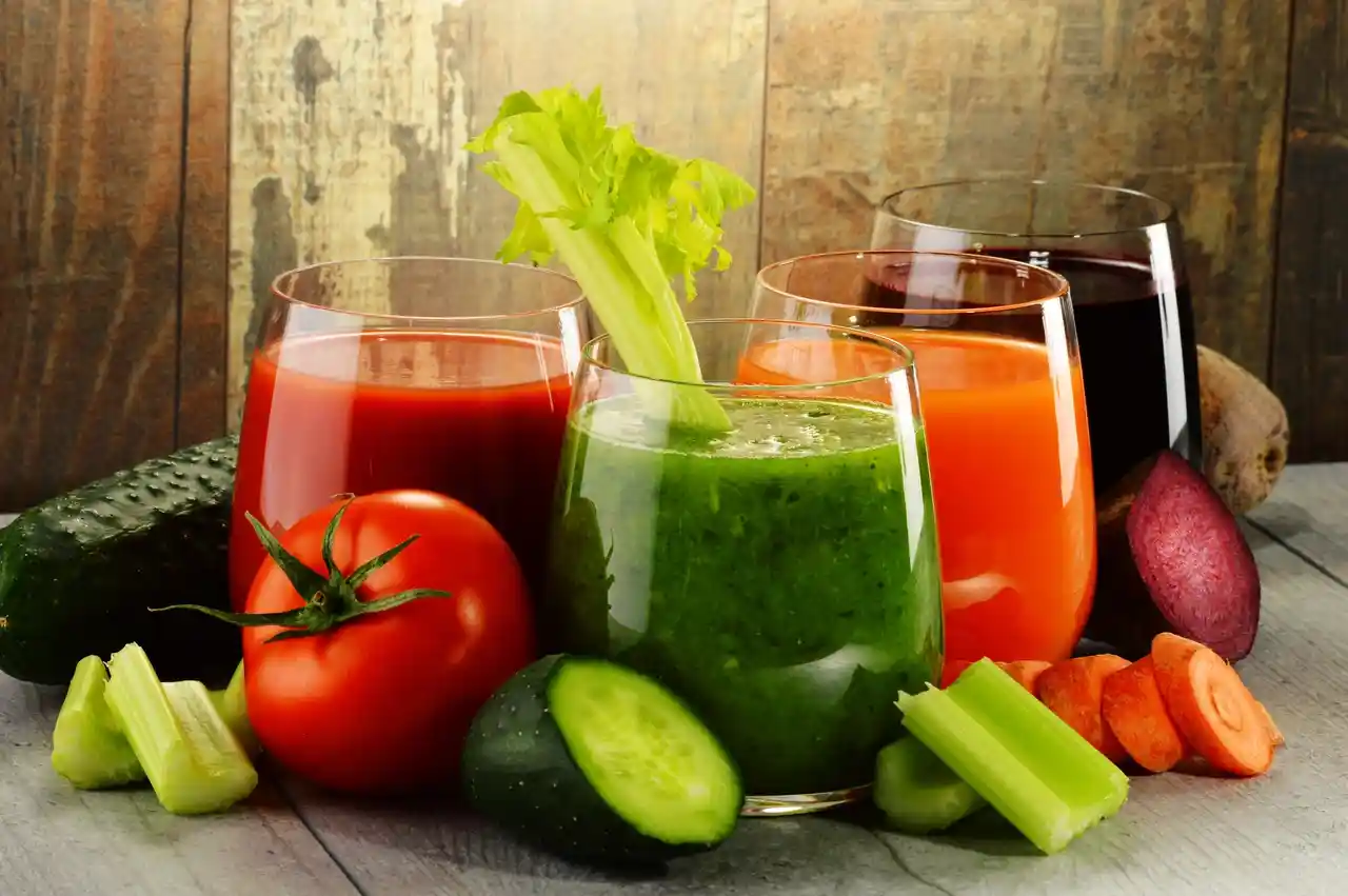 qt=q_30 (7) Four glass cups of vegetable juices on a wooden surface, surrounded by fresh tomatoes, cucumbers, celery, carrots, and beets.
